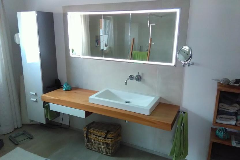 Ein helles Badezimmer mit maßgefertigter Holzwaschtischplatte, weißem Waschbecken, großem Spiegel mit Beleuchtung und passendem Hochschrank.