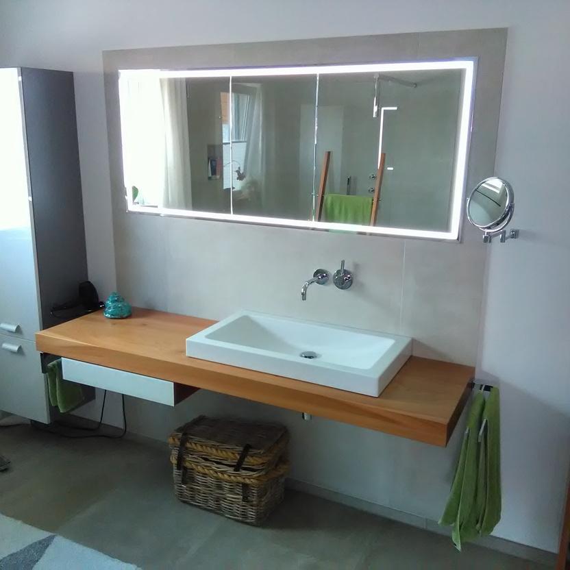 Ein helles Badezimmer mit maßgefertigter Holzwaschtischplatte, weißem Waschbecken, großem Spiegel mit Beleuchtung und passendem Hochschrank.