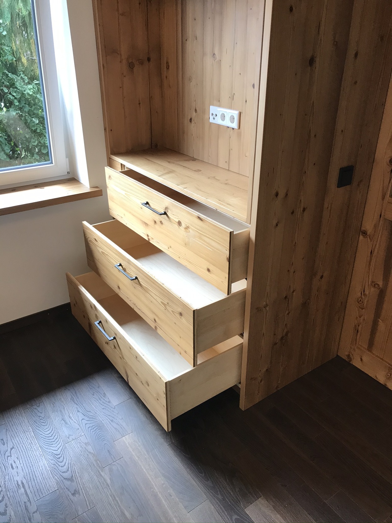 Maßgefertigte Holzschubladen in einer Ecke neben einem Fenster, hervorgehoben durch dunklen Holzboden.
