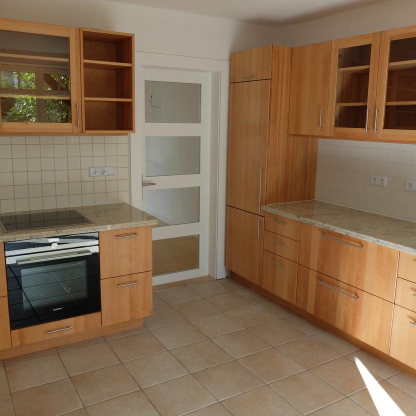 Eine Ecke einer Küche nach Maß mit hellen Holzschränken, integriertem Ofen und Kochfeld, Granitarbeitsplatte und weißen Fliesen.