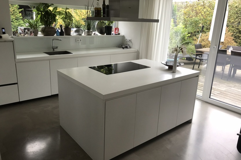 Eine schicke weiße Kücheninsel im Zentrum einer Küche nach Maß mit Blick auf einen Garten durch bodentiefe Fenster.