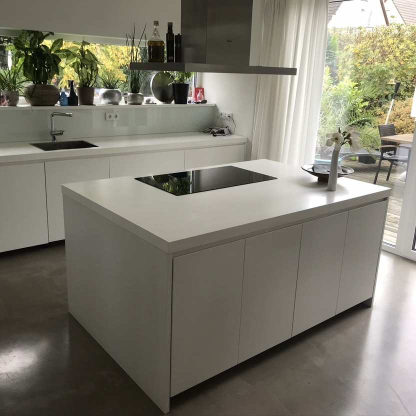 Eine schicke weiße Kücheninsel im Zentrum einer Küche nach Maß mit Blick auf einen Garten durch bodentiefe Fenster.