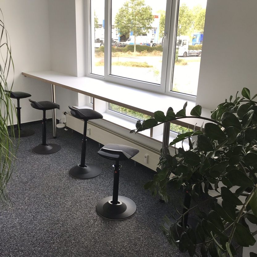 Gemütliche Büroecke mit einem langen Fenstertisch und modernen Hockern, umgeben von üppigen Grünpflanzen, die eine entspannte Atmosphäre schaffen.