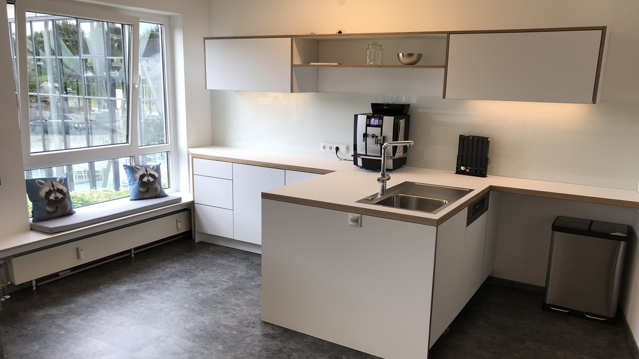 Helles, minimalistisch eingerichtetes Büro mit modernen Büromöbeln, darunter eine Küchenzeile mit Einbauschränken und Geräten.