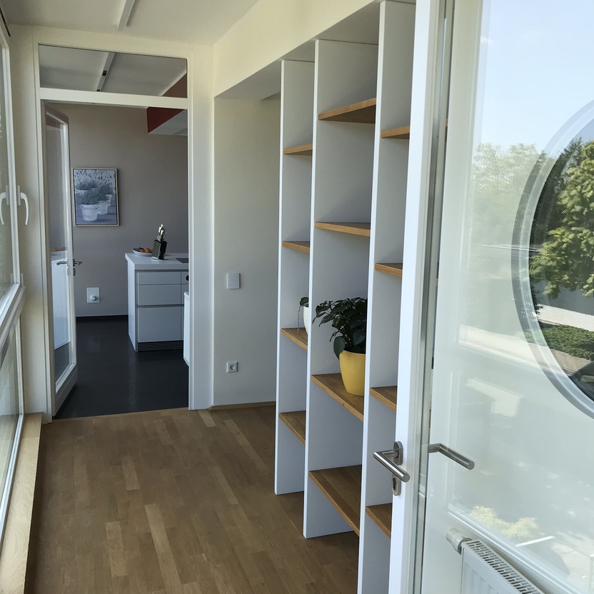 Helle, offene Raumgestaltung mit einem weißen Raumteiler, der Holzregale integriert, die zur Zonierung des Raumes und als dekorative Elemente dienen.